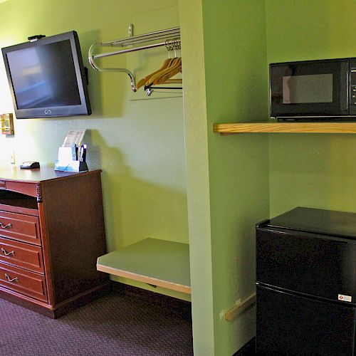 The image features a hotel room with a wall-mounted TV, wooden dresser, mirror, microwave, mini-fridge, and a shelf with hangers on a green wall.