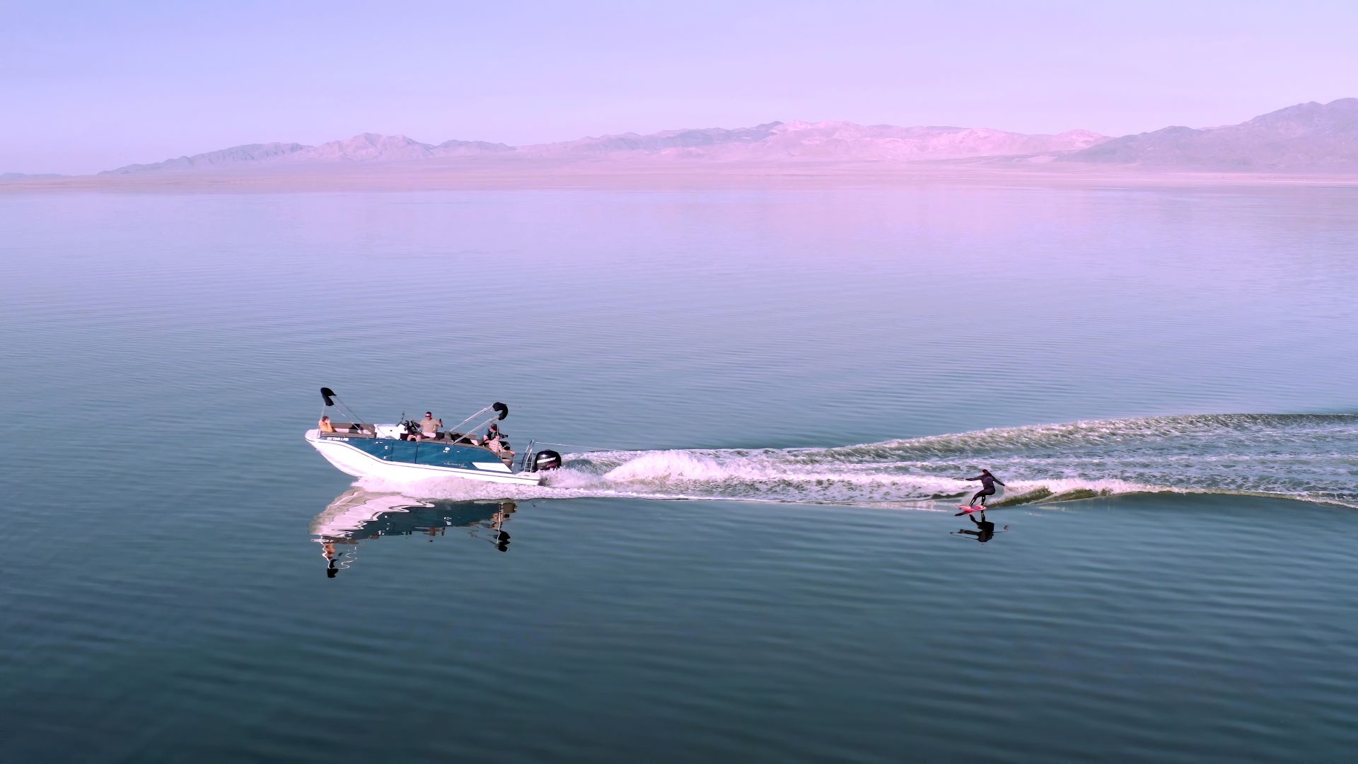 A speedboat pulls a person on a wakeboard across a calm body of water with a mountain range in the background under a clear sky.