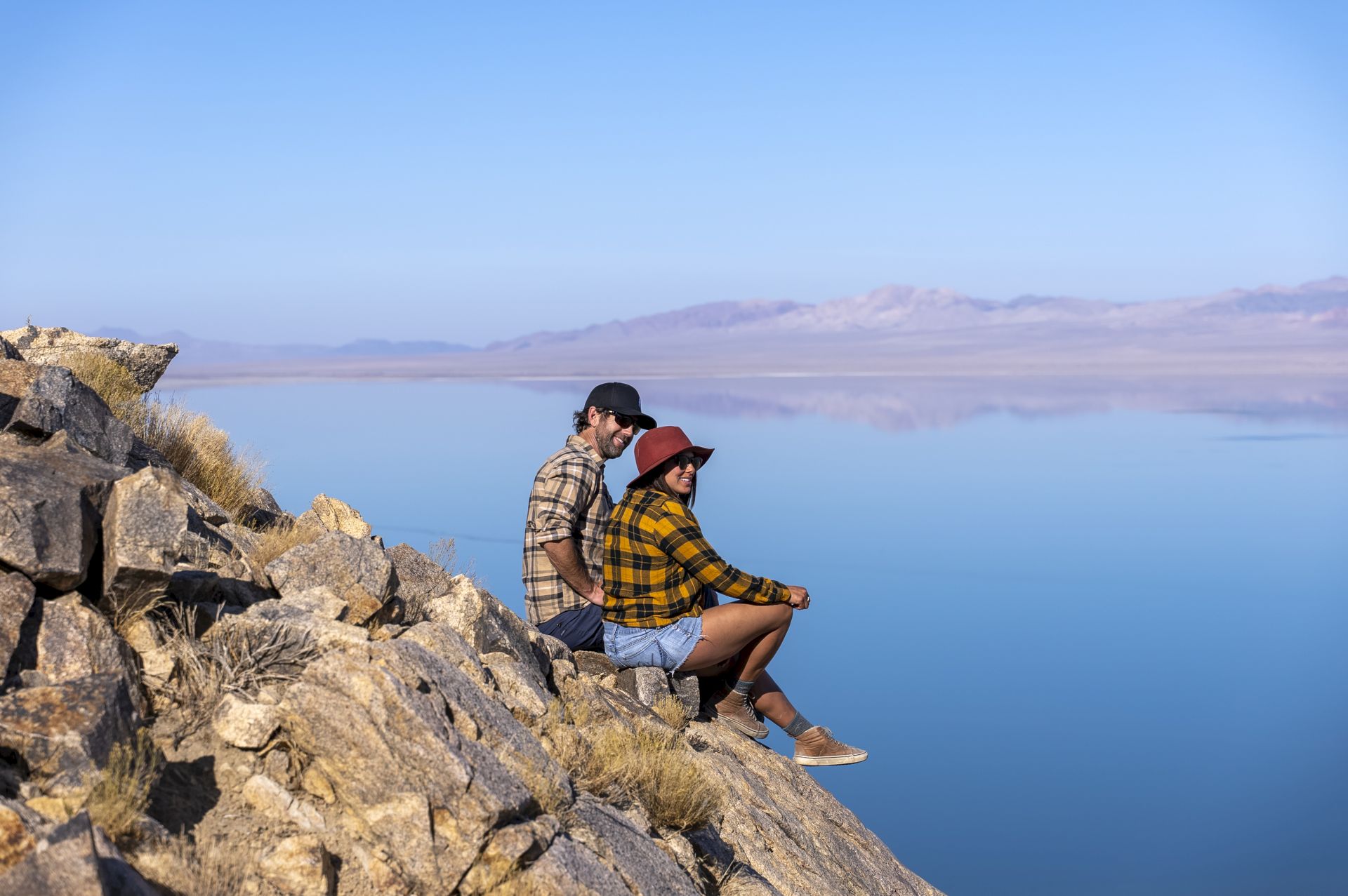 Two people are sitting on a rocky outcrop by a calm, expansive body of water, wearing casual outdoor attire.
