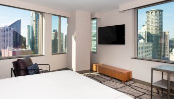 A modern hotel room with large windows offering a city view, a bed, armchair, TV on the wall, and a console table.