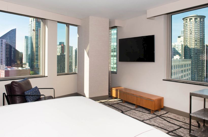 A modern hotel room with large windows offering a city view, a bed, armchair, TV on the wall, and a console table.