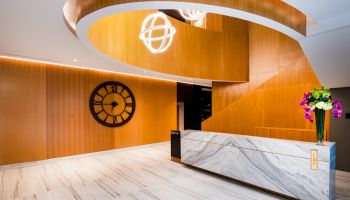 A modern lobby features a curved wooden staircase, marble reception desk with flowers, a large wall clock, and contemporary lighting fixtures.
