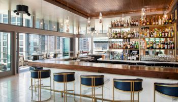 The image shows a modern bar with stylish seating, a well-stocked liquor shelf, and large windows offering a view of a cityscape.