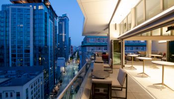A modern outdoor patio on a high-rise building at dusk, featuring tables and chairs, overlooking a cityscape with tall buildings and a street below.