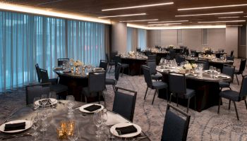 A banquet hall is elegantly set up with round tables, black chairs, and neatly arranged table settings, featuring white flowers and candles.