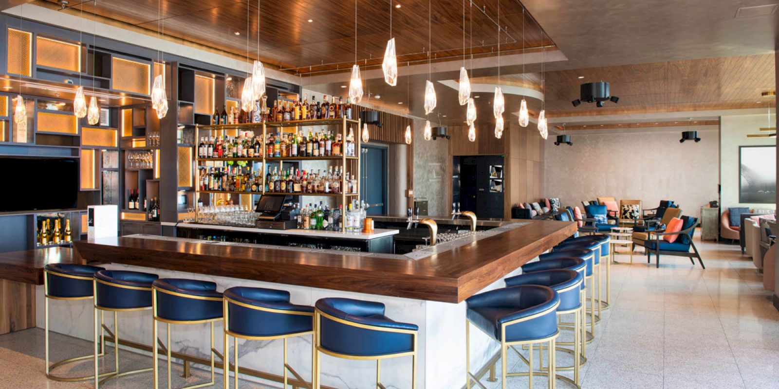 The image shows a modern indoor bar area with a long wooden bar counter, blue bar stools, shelves of alcohol bottles, and pendant lights.
