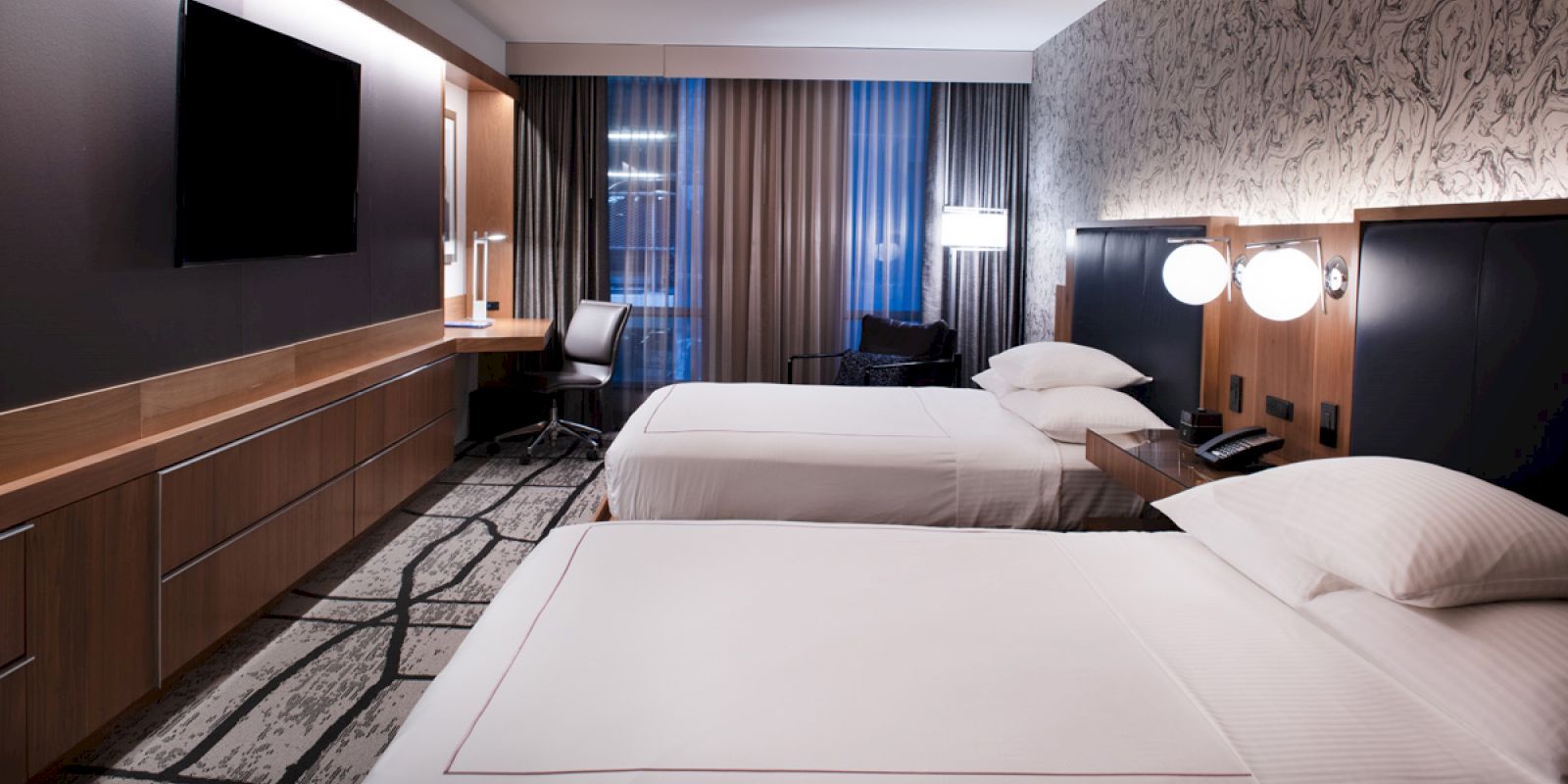 A modern hotel room with two beds, a TV, a work desk, and large windows with curtains.