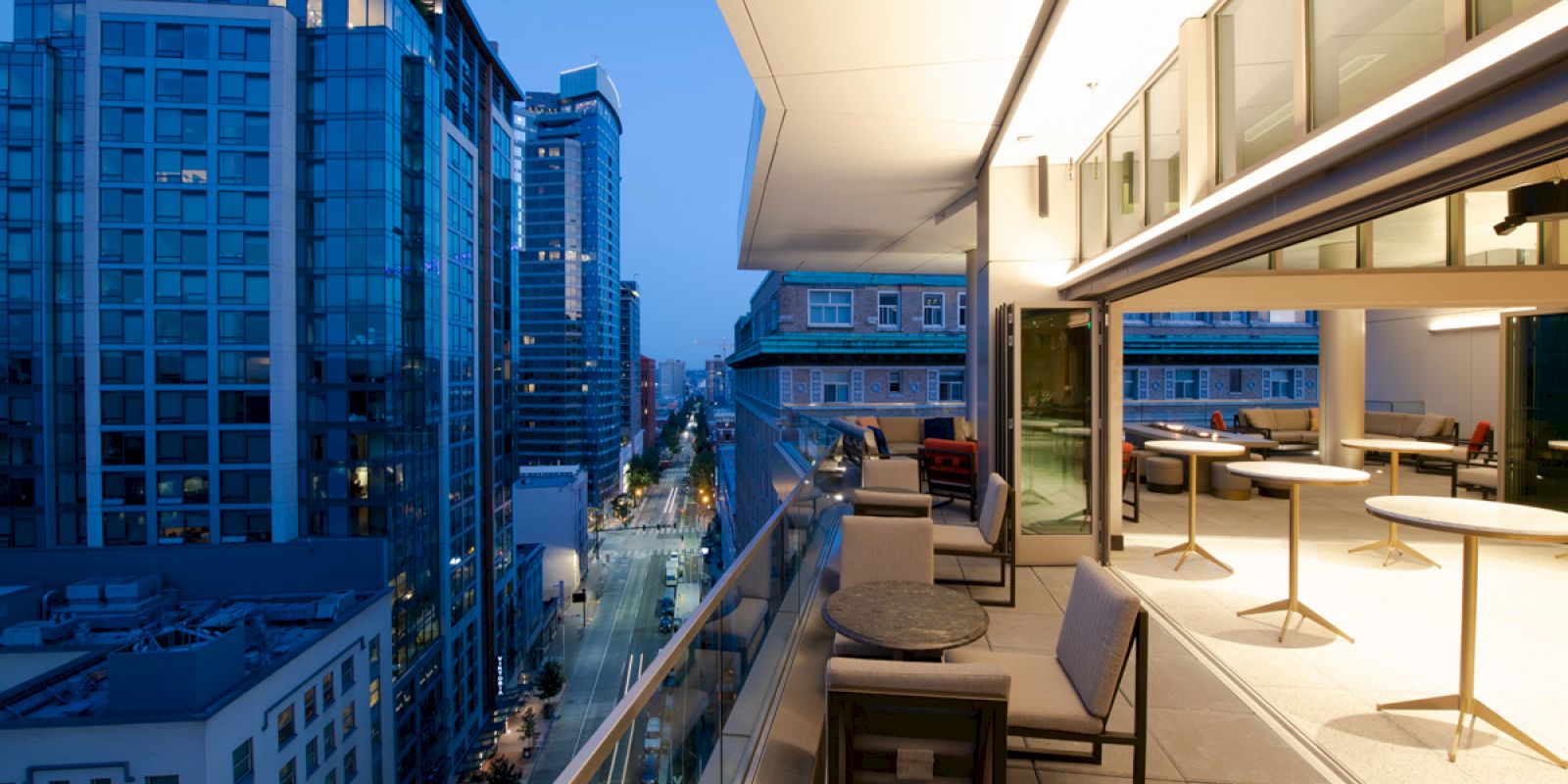 A modern cityscape at dusk with tall buildings and a rooftop terrace featuring tables and chairs overlooking the street below.