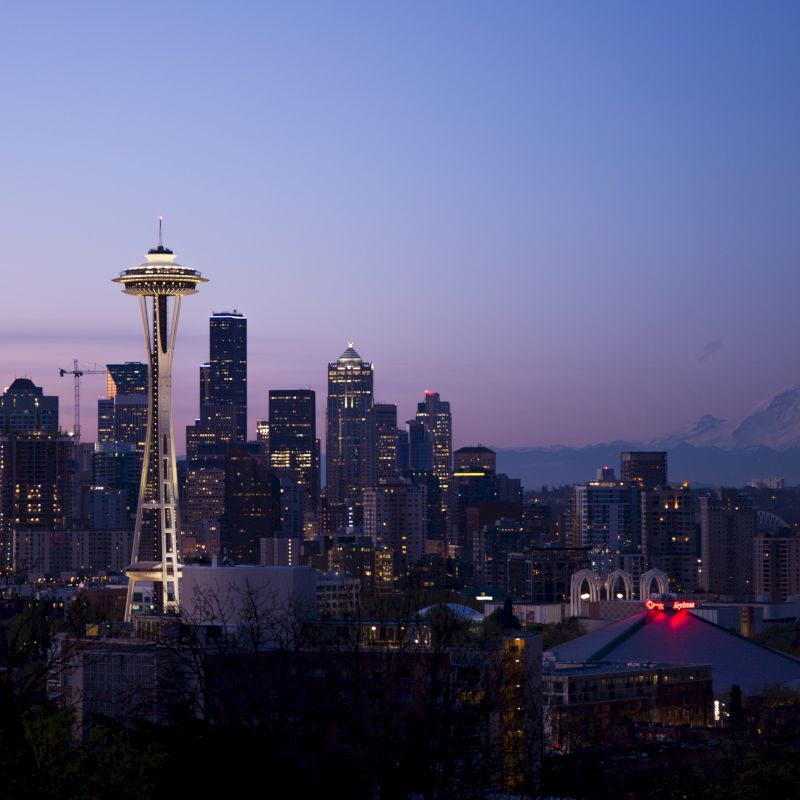 This image shows a cityscape at dusk, featuring the Space Needle in Seattle with Mount Rainier in the background and surrounding buildings.