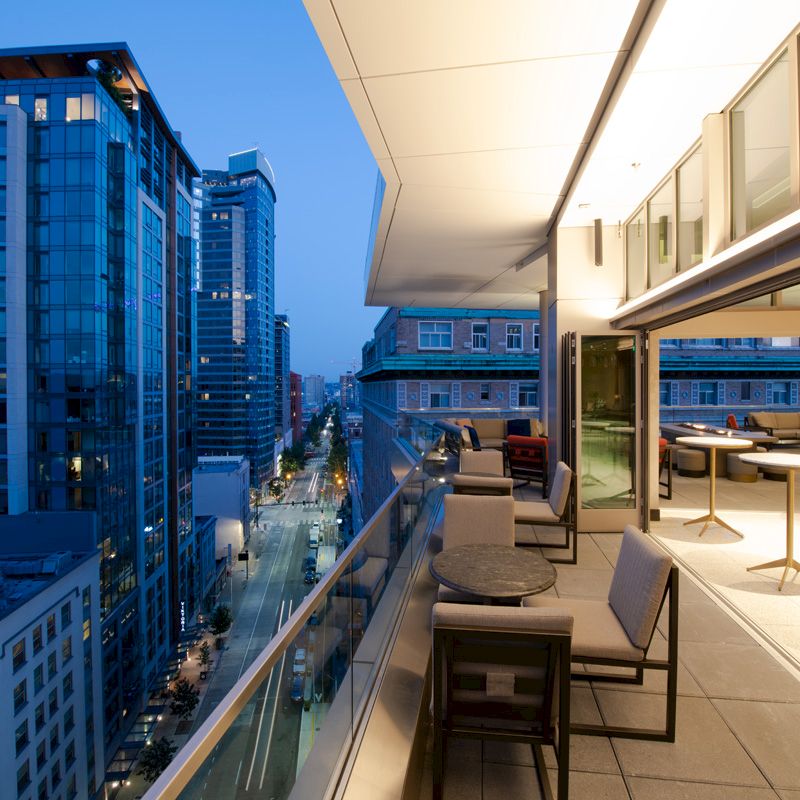 A modern cityscape is viewed from a sleek balcony with seating and tables, high-rise buildings are seen along a lit street during twilight.