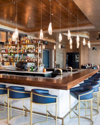 The image shows a modern bar with high chairs, a well-stocked liquor shelf, hanging lights, and a seating area in the background.