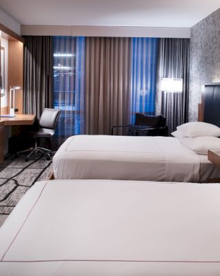 A modern hotel room with two double beds, a TV, desk, and large window with curtains. The room has stylish decor and ambient lighting.