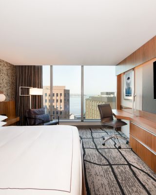 A modern hotel room with a king-sized bed, a large flat-screen TV, a desk, and a chair, plus a window providing a view of city buildings.