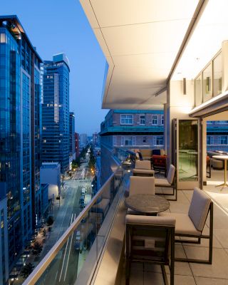 A modern balcony with seating and tables overlooking a city street with tall buildings at dusk.
