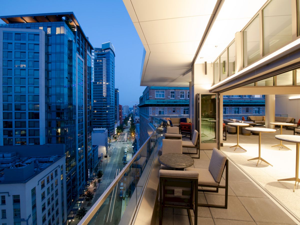 A modern cityscape with tall buildings at dusk, featuring an outdoor balcony area with seating and tables on the right side.
