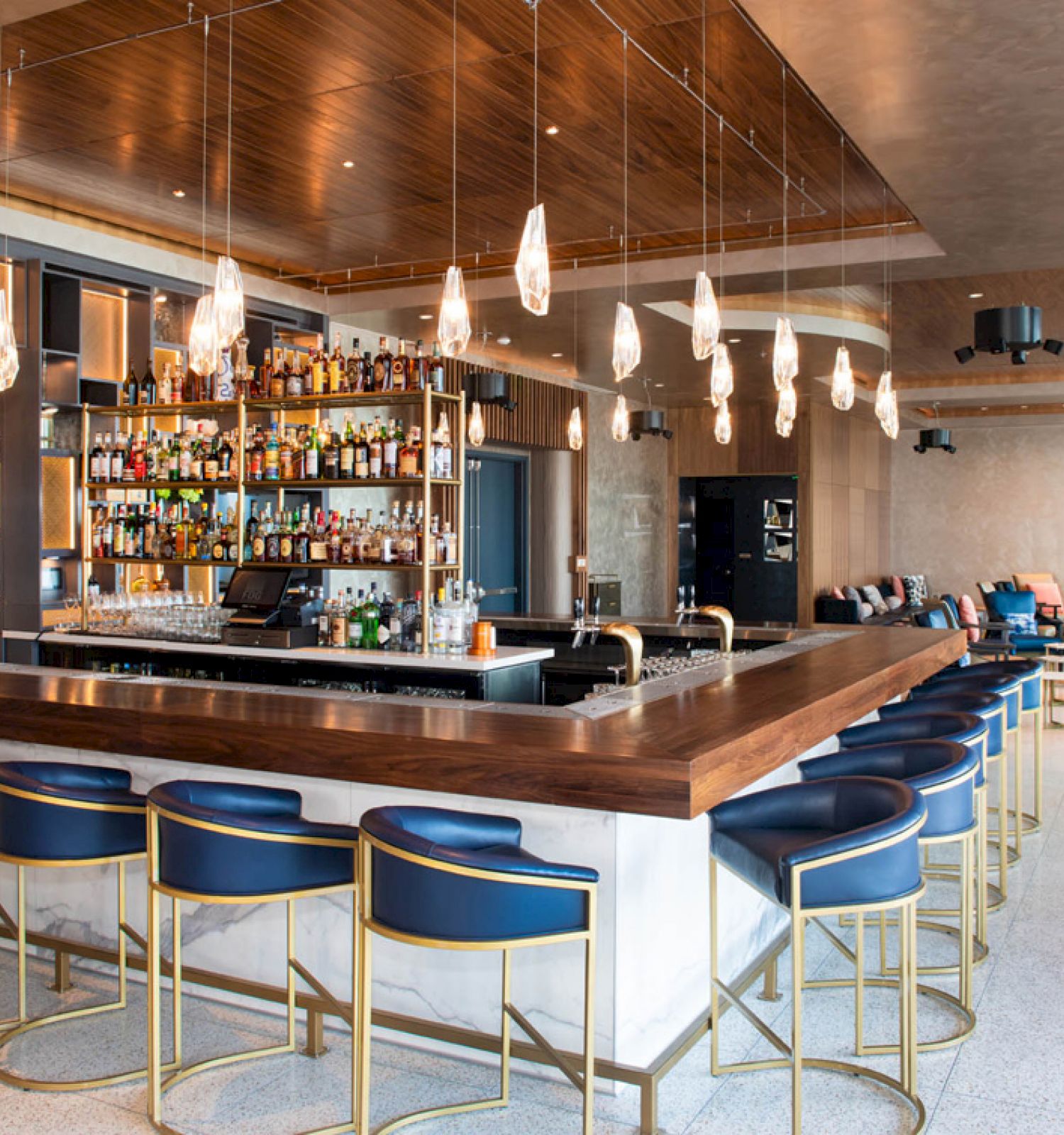 This image shows a modern bar with blue chairs, a wooden countertop, various bottles on shelves, hanging lights, and a seating area in the back.