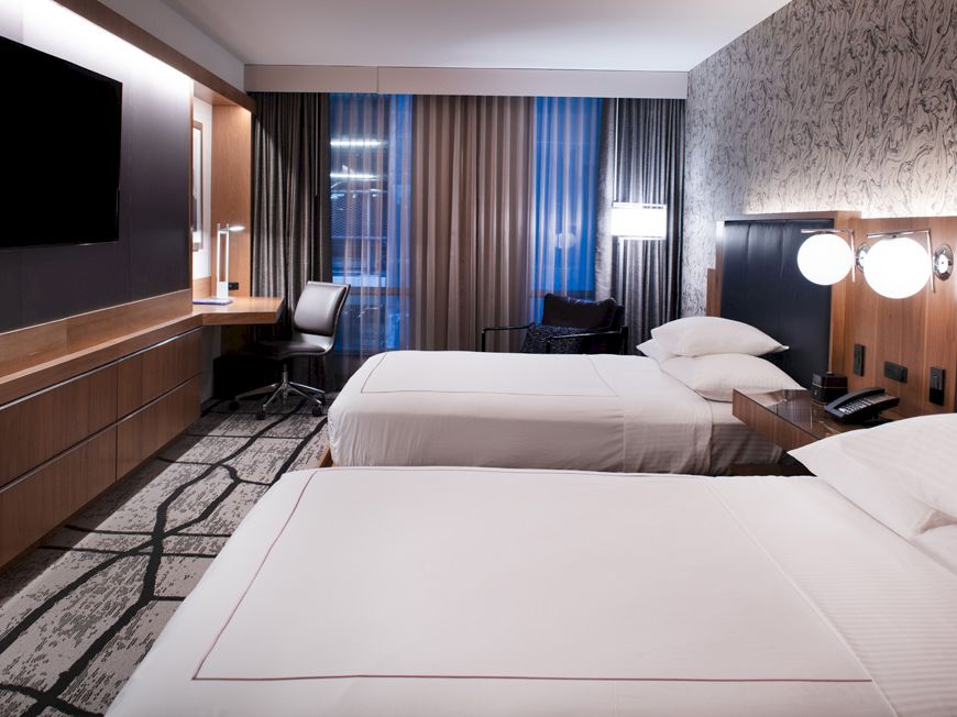 The image shows a modern hotel room with two beds, a television, a desk with a chair, and large windows with curtains, in a minimalist style.