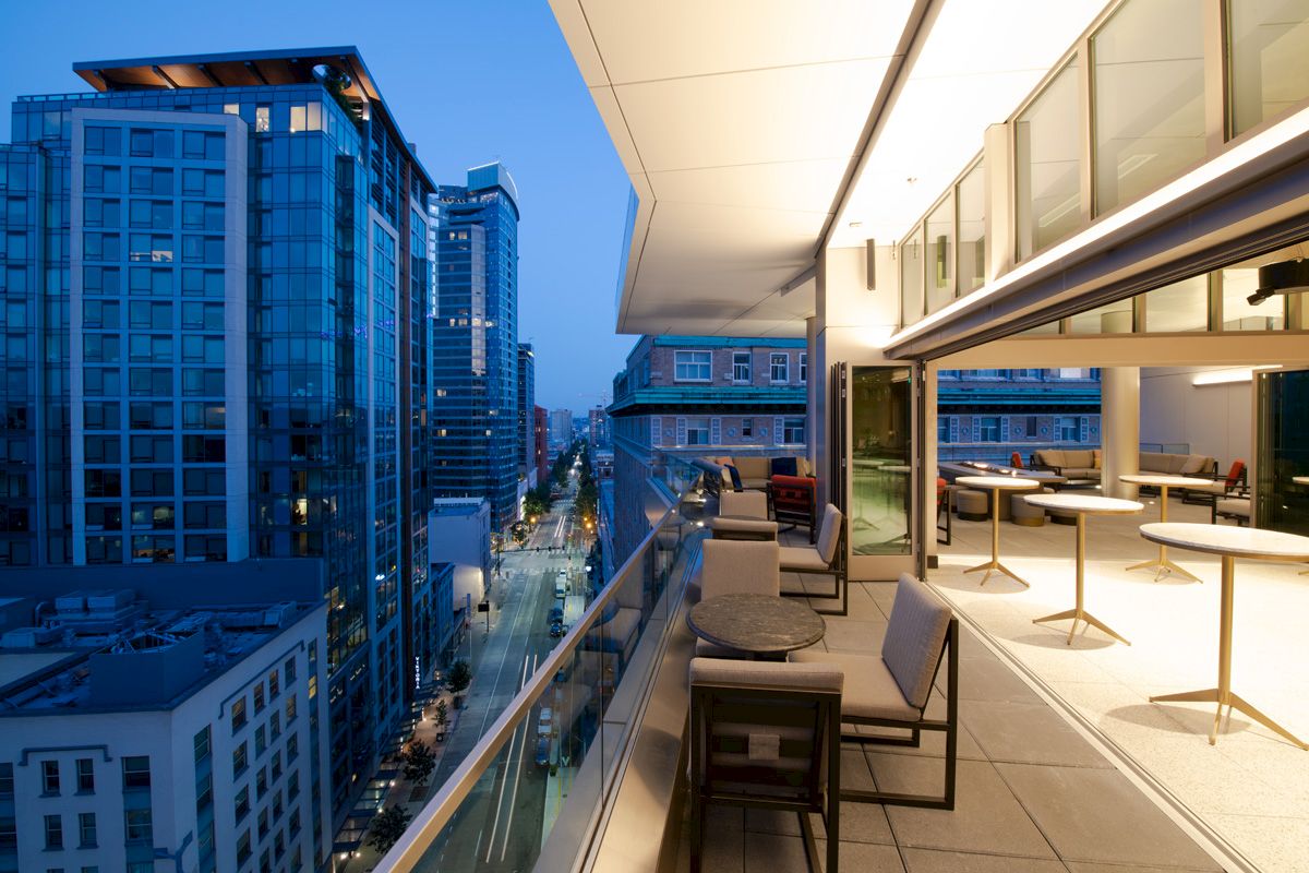 Modern city view from a high-rise balcony at dusk, featuring outdoor seating and tables, surrounded by tall buildings and illuminated street below.