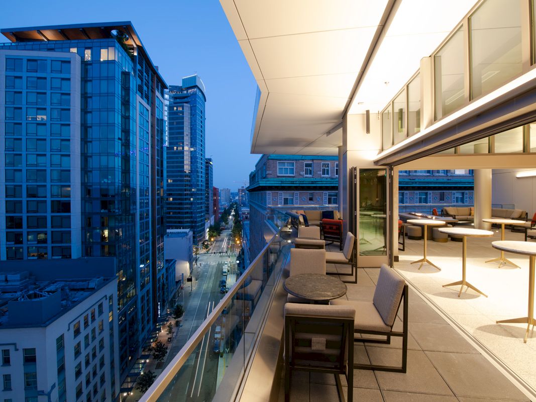 Modern city view from a high-rise balcony at dusk, featuring outdoor seating and tables, surrounded by tall buildings and illuminated street below.