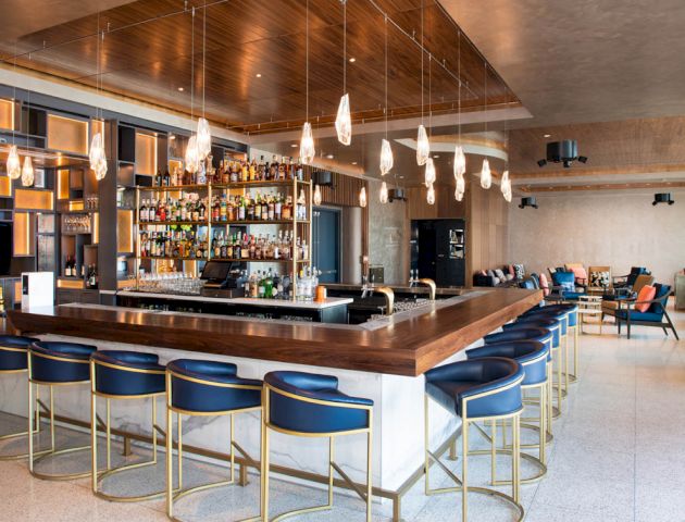 A modern bar with a well-stocked liquor shelf, blue bar stools, stylish lighting, and a cozy seating area in the background.