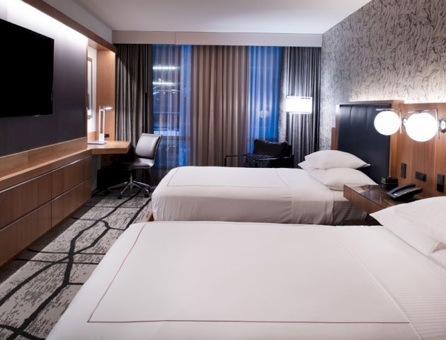A modern hotel room features two beds, a flat-screen TV, a desk with a chair, bedside lamps, and large windows with curtains.
