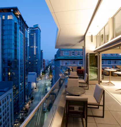 A modern cityscape with tall buildings at dusk, viewed from a spacious balcony or patio area featuring tables, chairs, and soft lighting.