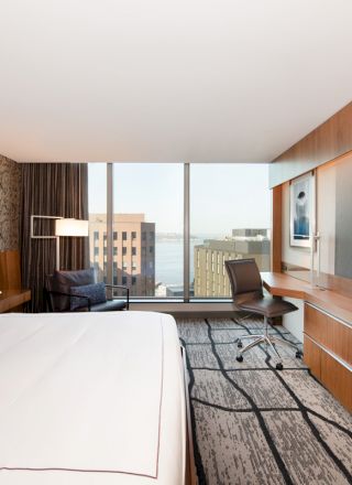 The image shows a modern hotel room with a large bed, desk, chair, and wall-mounted TV. The room has a large window with a city view.