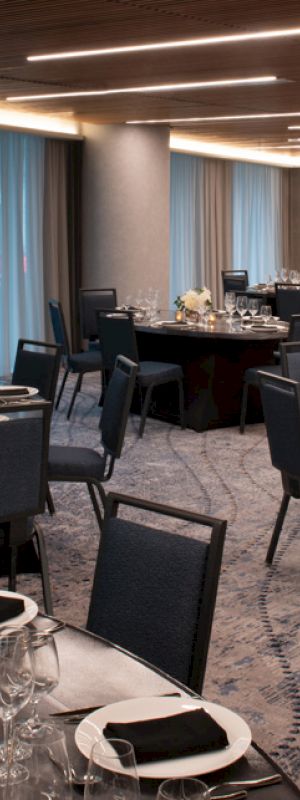 A spacious, elegant dining room is set for an event, featuring round tables with black tablecloths and neatly arranged chairs.