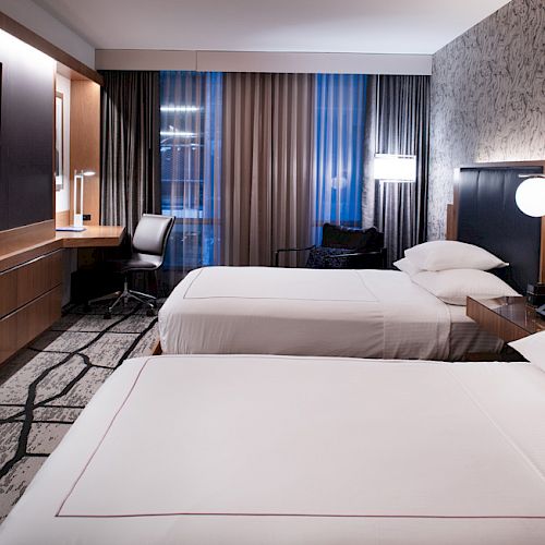 The image shows a modern hotel room with two beds, a desk, a chair, a large TV, and stylish decor, featuring a patterned accent wall and clean lines.