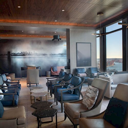 A lounge area featuring cushioned seats, tables, a cityscape mural, and large windows offering a view of a distant Ferris wheel and waterfront.
