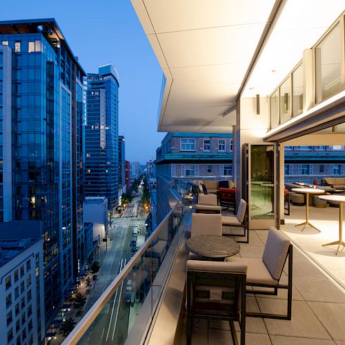 A modern balcony with tables and chairs overlooking a city street filled with tall buildings at dusk. The interior is well-lit and inviting.