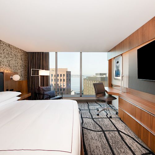 The image shows a modern hotel room with a large bed, a desk, a chair, a flat-screen TV, and a window with a view of buildings and water.