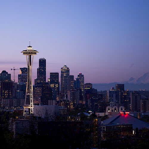 The image shows the Seattle skyline during twilight, featuring the Space Needle prominently. Mount Rainier is visible in the background.