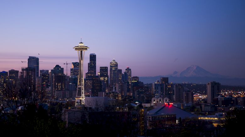 The image shows the Seattle skyline with the Space Needle prominently lit against a twilight sky and Mount Rainier in the background.