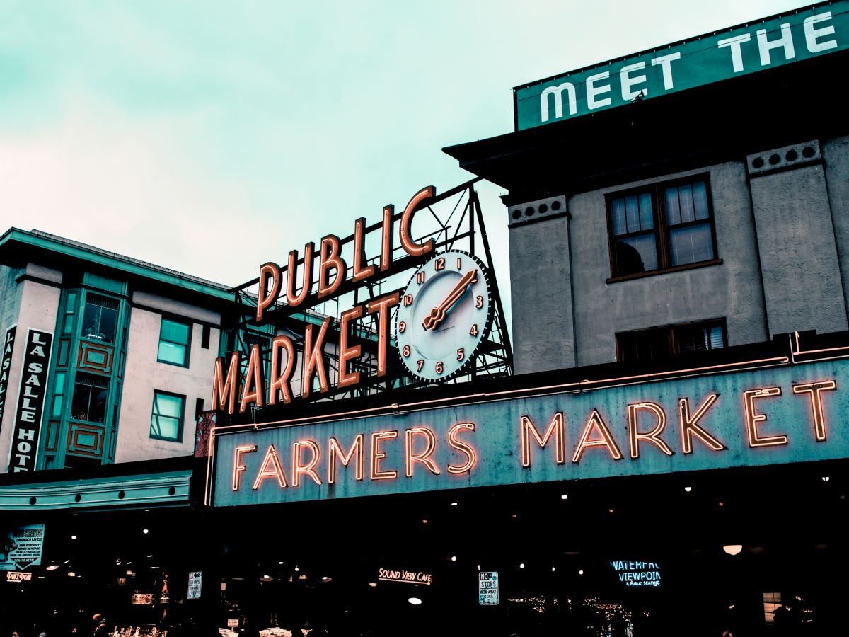 The image shows a vibrant sign for a "Public Market" and "Farmers Market" with a clock, located in a bustling urban area, under a cloudy sky.