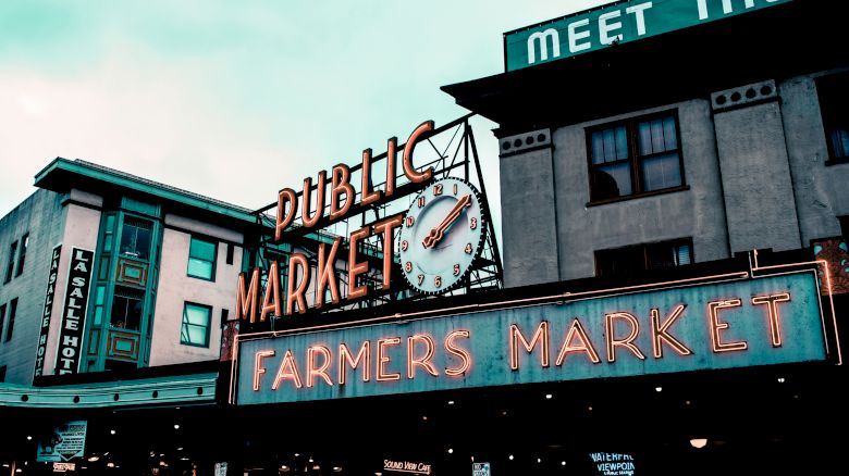 The image shows a vibrant sign for a "Public Market" and "Farmers Market" with a clock, located in a bustling urban area, under a cloudy sky.