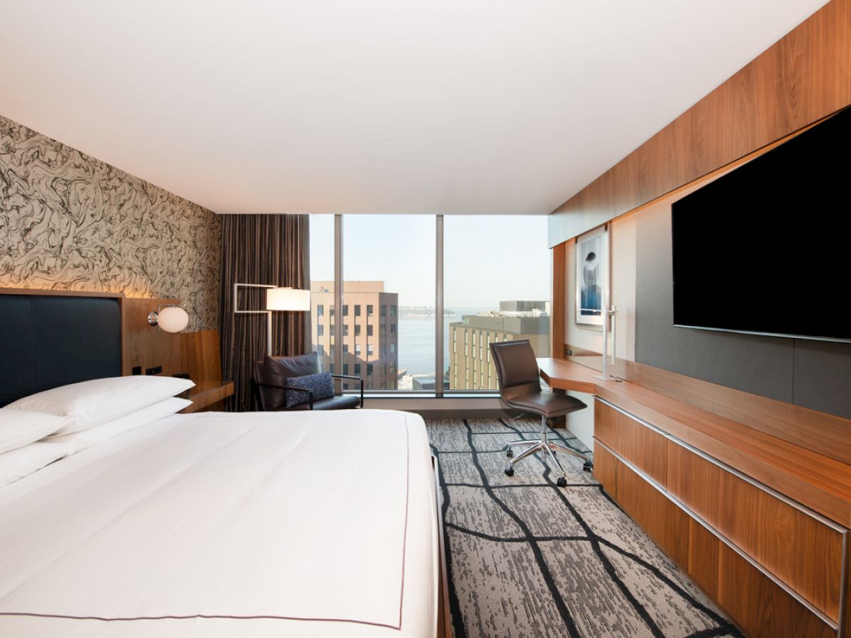 A modern hotel room with a large bed, a wall-mounted TV, a work desk, and a view of city buildings through floor-to-ceiling windows.