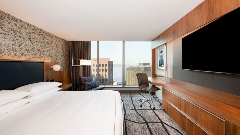 A modern hotel room with a large bed, a wall-mounted TV, a work desk, and a view of city buildings through floor-to-ceiling windows.