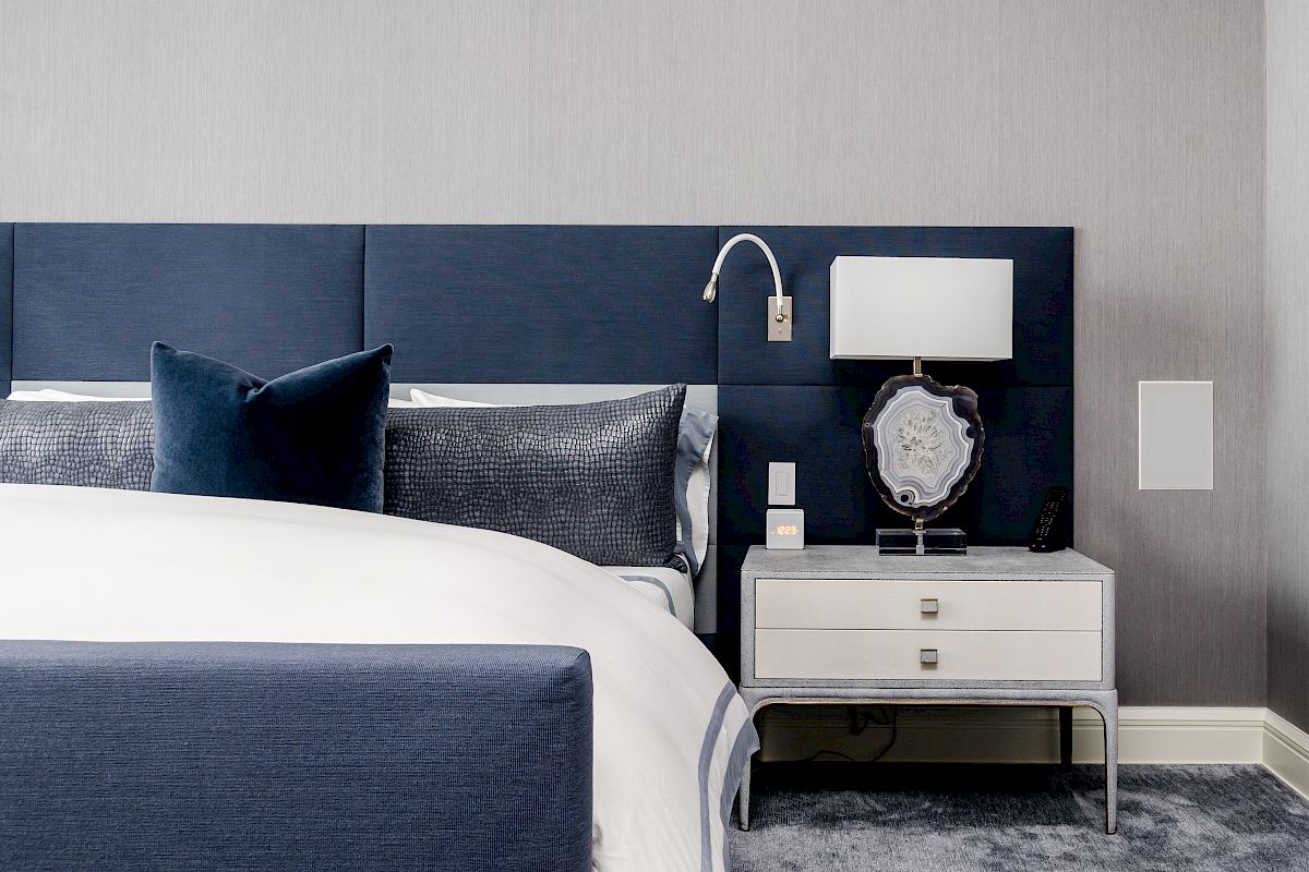 Modern bedroom with blue headboard, white bed linens, gray pillows, and a white nightstand with a lamp and decorative item on top.