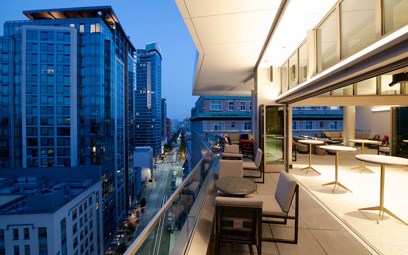 A modern balcony with seating overlooks a cityscape filled with tall buildings at dusk, featuring large windows and a city street below.