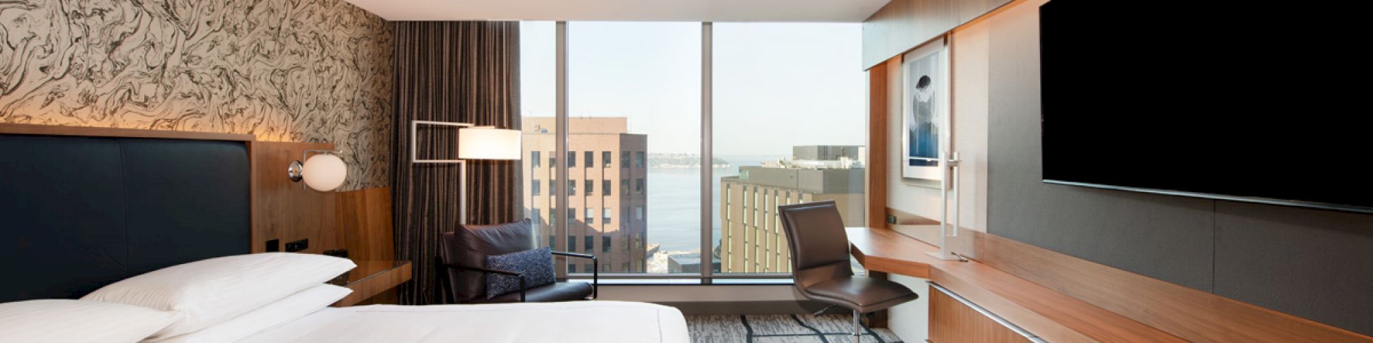 This image shows a modern hotel room with a large bed, a flat-screen TV, a desk, and a city view through large windows.