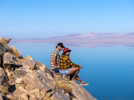 Two people sit on a rocky ledge overlooking a serene lake and distant mountains under a clear blue sky, both wearing plaid outfits and hats.